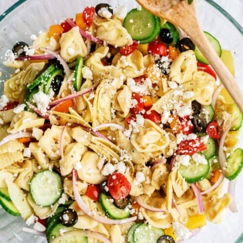 A glass bowl of tortellini pasta salad with cucumbers, cherry tomatoes, artichoke hearts, olives, red onion, and feta cheese, with a wooden spoon.