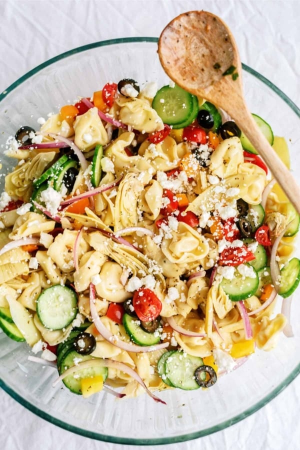 A glass bowl of tortellini pasta salad with cucumbers, cherry tomatoes, artichoke hearts, olives, red onion, and feta cheese, with a wooden spoon.