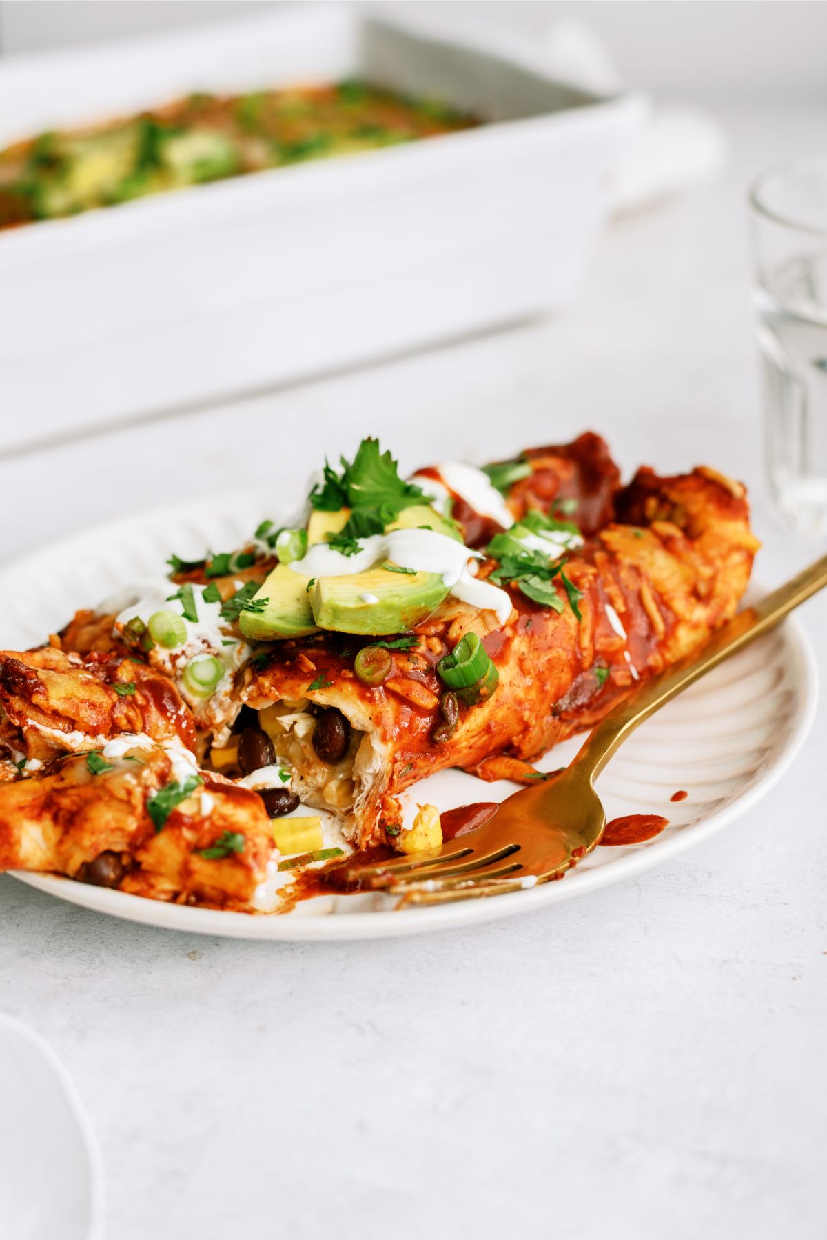 Two Chicken Fiesta Enchiladas on a plate with toppings, a pan of Chicken Fiesta Enchiladas in the background