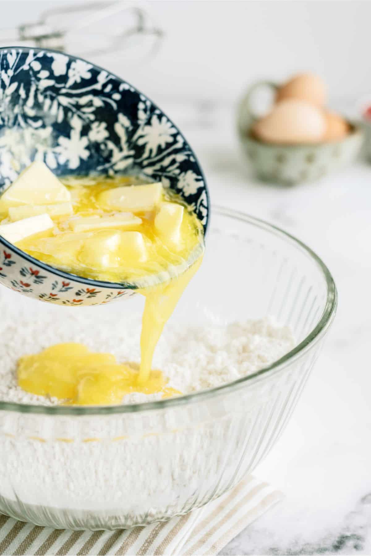 A mixing bowl with flour and eggs, with a bowl pouring melted butter into the bowl.