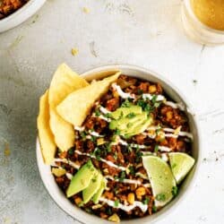 A bowl of Mexican rice topped with avocado slices, lime wedges, chopped herbs, tortilla chips, and drizzled with crema, next to a glass of light-colored beverage.