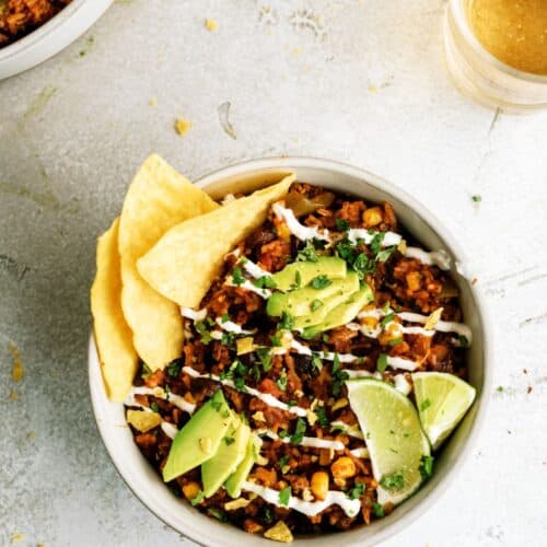 A bowl of Mexican rice topped with avocado slices, lime wedges, chopped herbs, tortilla chips, and drizzled with crema, next to a glass of light-colored beverage.