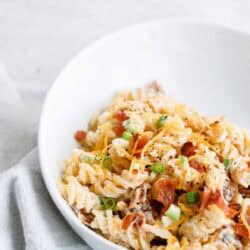 A white bowl of pasta salad with rotini, cheese, diced tomatoes, green onions, and bacon bits on a light gray cloth.