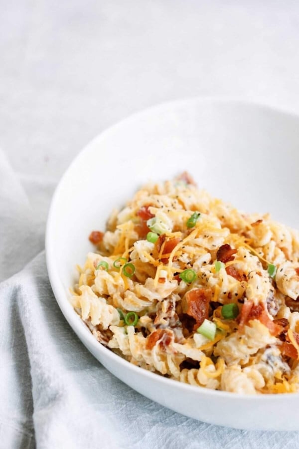A white bowl of pasta salad with rotini, cheese, diced tomatoes, green onions, and bacon bits on a light gray cloth.