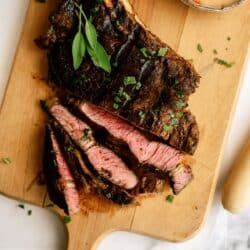 A sliced, medium-rare steak with herbs on a wooden cutting board, next to a small bowl of creamy sauce.