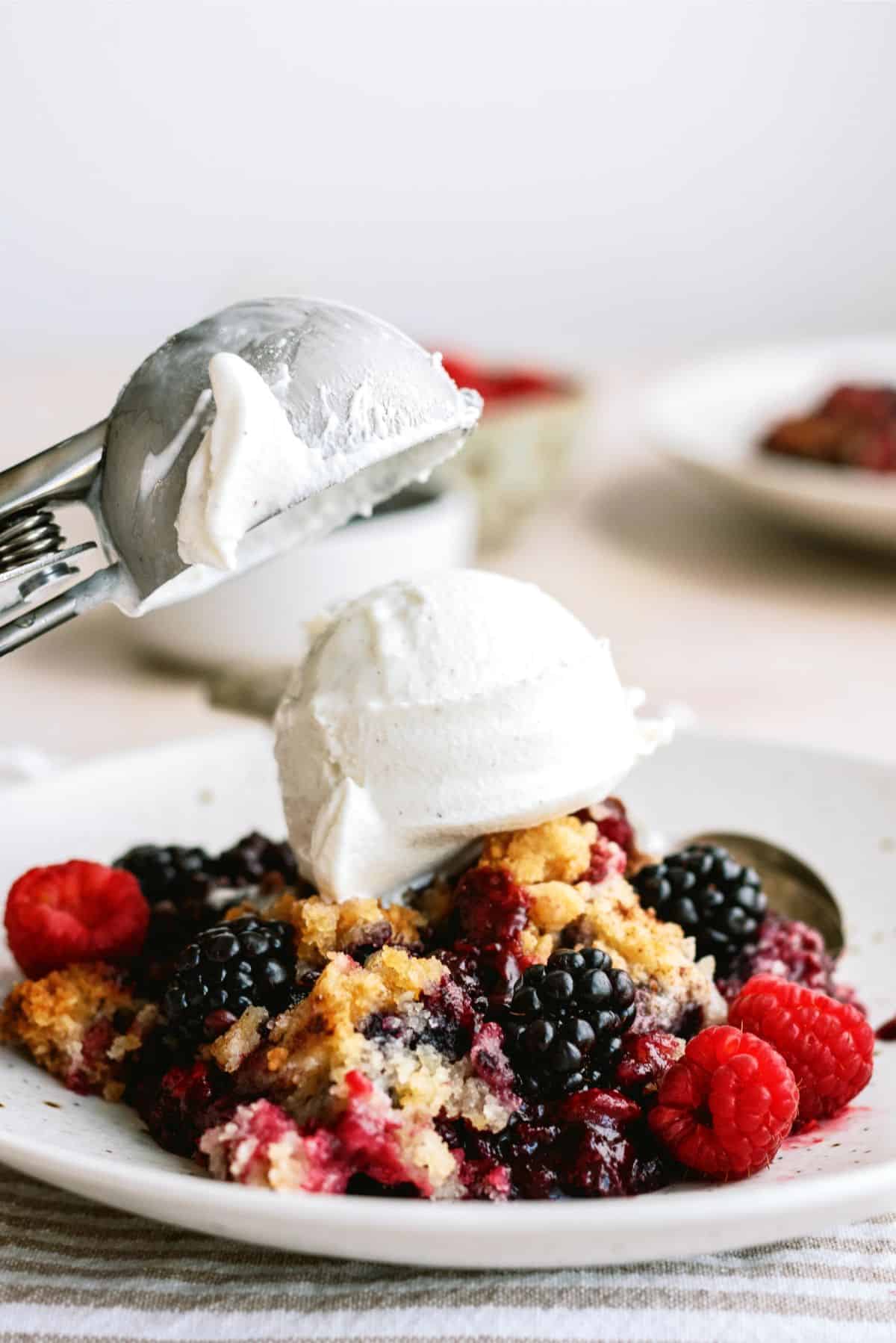 A scoop of vanilla ice cream is placed on top of a mixed berry cobbler with blackberries and raspberries on a white plate.