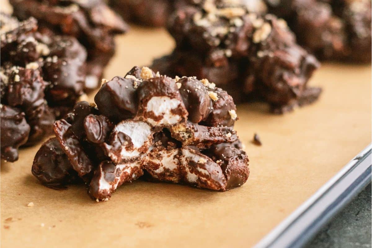 S’mores Cluster cut in half to show the inside on a baking sheet lined with parchment paper. Surrounded by other S’mores Clusters on the baking sheet.