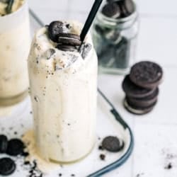 A tall glass of cookies and cream milkshake with a black straw, topped with crumbled chocolate sandwich cookies, on a tray with scattered cookies and a jar of cookies in the background.