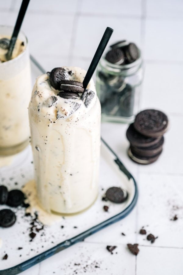 A tall glass of cookies and cream milkshake with a black straw, topped with crumbled chocolate sandwich cookies, on a tray with scattered cookies and a jar of cookies in the background.