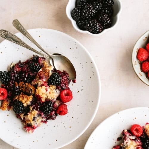 Plates of berry crumble with fresh raspberries and blackberries, served with spoons, beside small bowls of berries on a light surface.