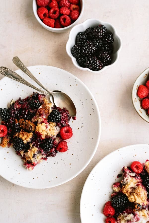 Plates of berry crumble with fresh raspberries and blackberries, served with spoons, beside small bowls of berries on a light surface.