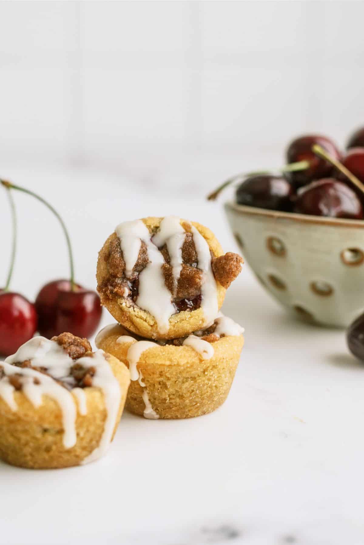 Three mini muffins with white icing are stacked in the foreground, with fresh cherries and a bowl of cherries in the background.