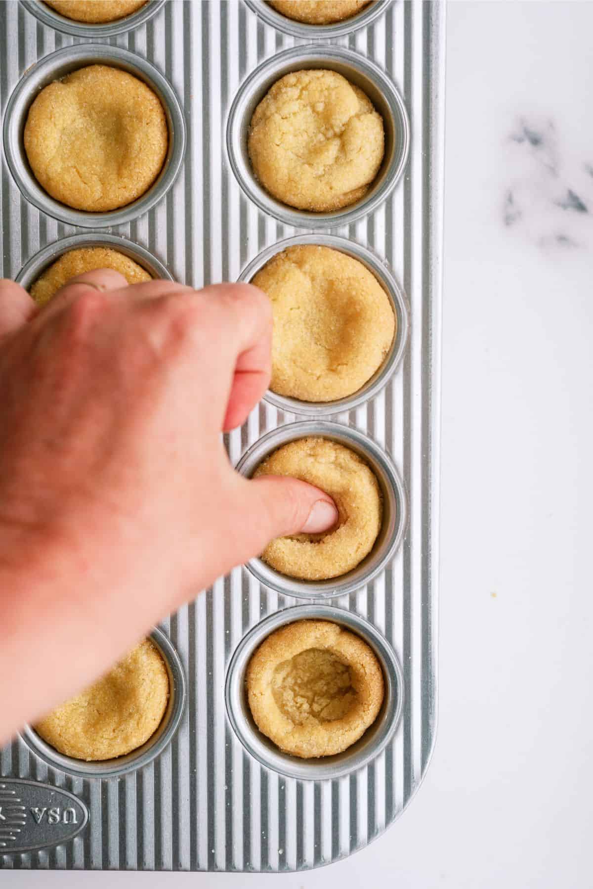 Pressing a thumb indent into a freshly baked sugar cookies in muffin tin.