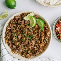 A bowl of Copycat Chipotle Barbacoa topped with 3 lime wedges with a bowl of white rice on the side.