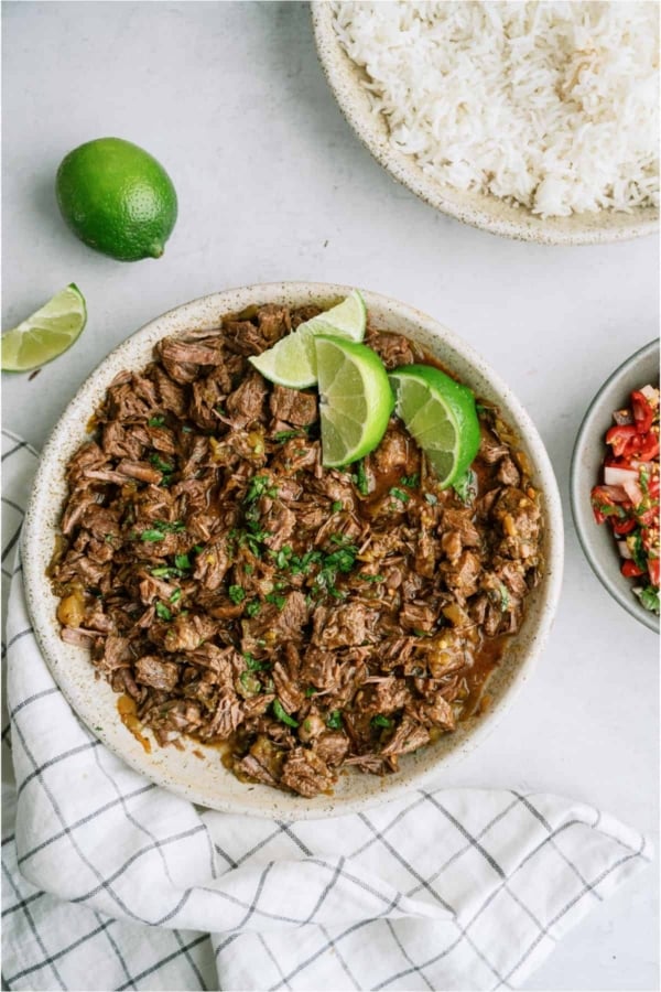 A bowl of Copycat Chipotle Barbacoa topped with 3 lime wedges with a bowl of white rice on the side.