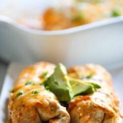 A close-up of a plated enchilada topped with melted cheese and avocado slices. In the background, a baking dish with more enchiladas is visible.