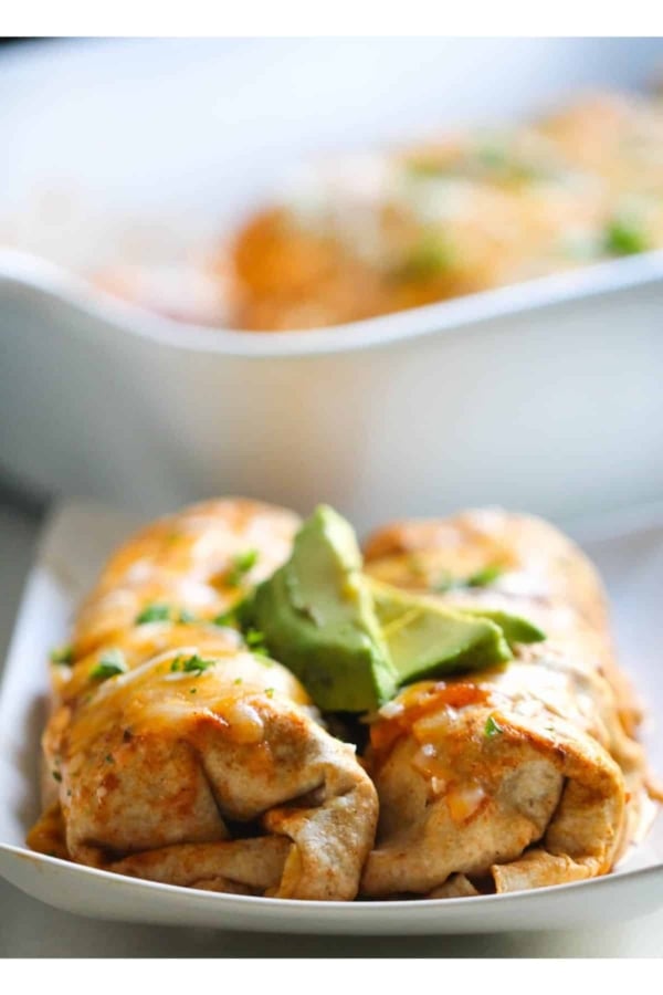A close-up of a plated enchilada topped with melted cheese and avocado slices. In the background, a baking dish with more enchiladas is visible.