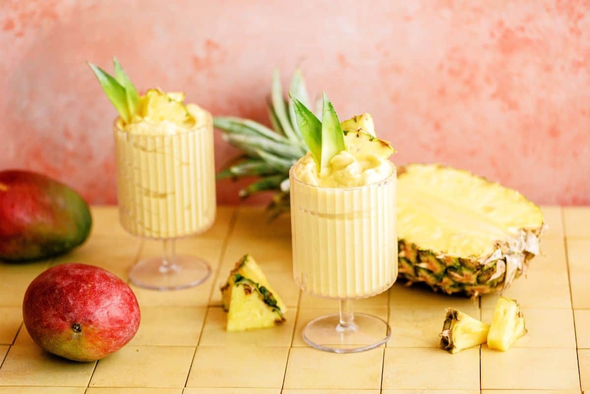 2 glasses of Mango and Pineapple Dole Whip surrounded by pineapple and mangpes