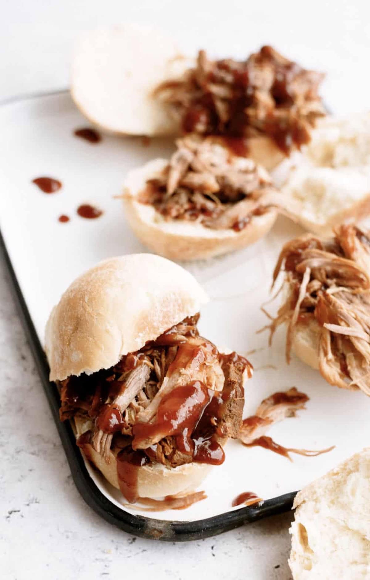 Pulled pork sandwiches on a white tray, topped with barbecue sauce, with sandwich buns and some sauce drips visible.