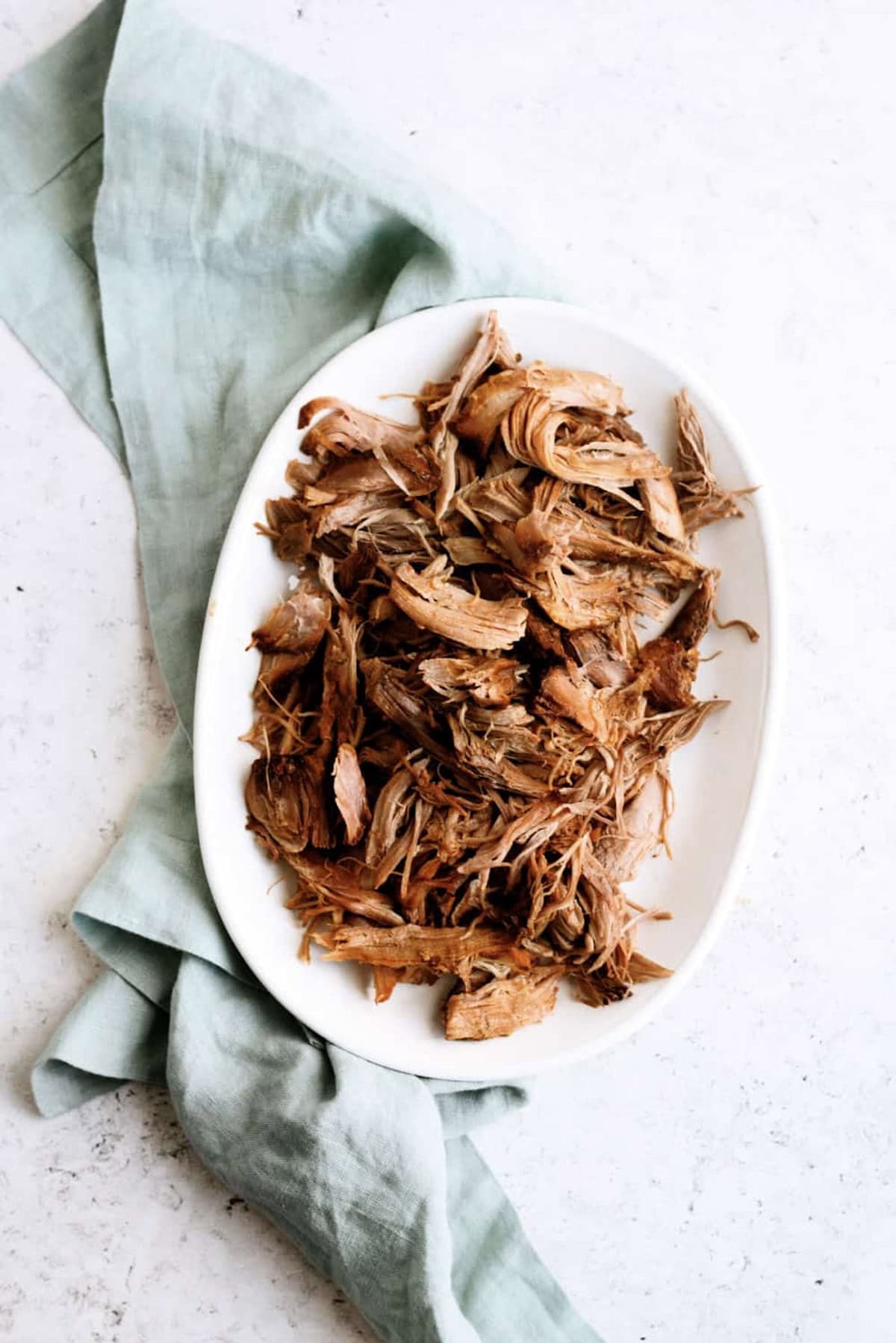 Shredded cooked meat served on a white oval plate, placed on top of a light blue cloth napkin on a white surface.