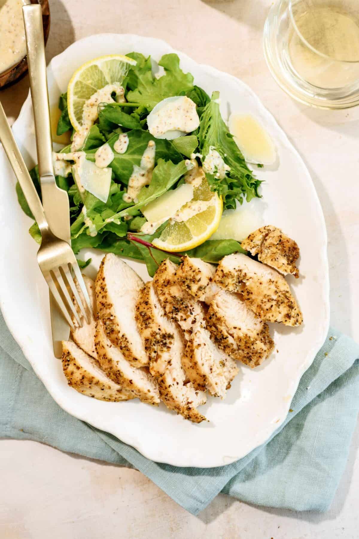 Sliced seasoned chicken breast with a green salad, lemon wedges, and dressing on a white plate, with a fork and knife on the side.