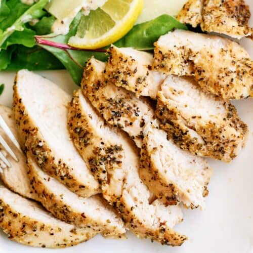 Sliced seasoned chicken breast served with fresh greens, a lemon wedge, and salad dressing on a white plate.