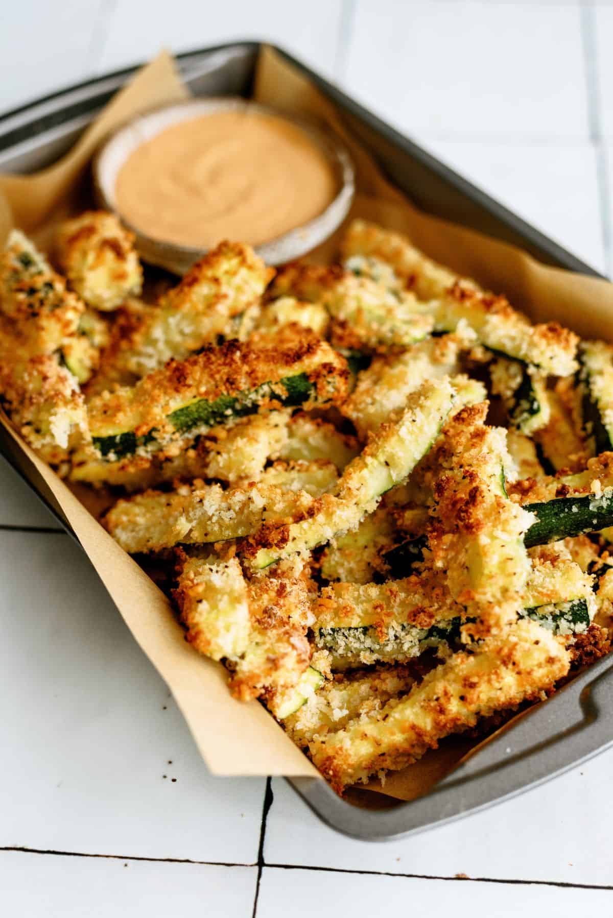 A sheet pan of Crispy Zucchini Fries with a cup of creamy BBQ Sauce