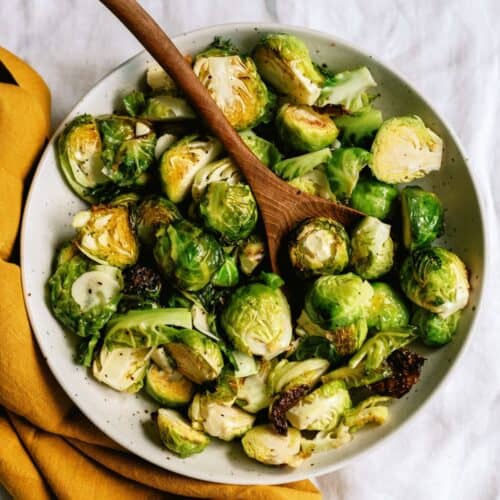 A bowl of roasted Brussels sprouts with a wooden spoon, placed on a white surface next to a mustard-yellow cloth napkin.