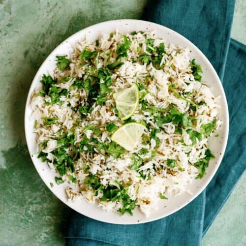 A white plate of herbed rice garnished with chopped herbs and lime wedges, placed on a dark green napkin against a textured green background.