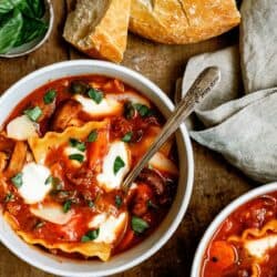 Two bowls of lasagna soup with melted cheese and basil sit on a wooden table next to a loaf of crusty bread and a small dish of fresh basil leaves.