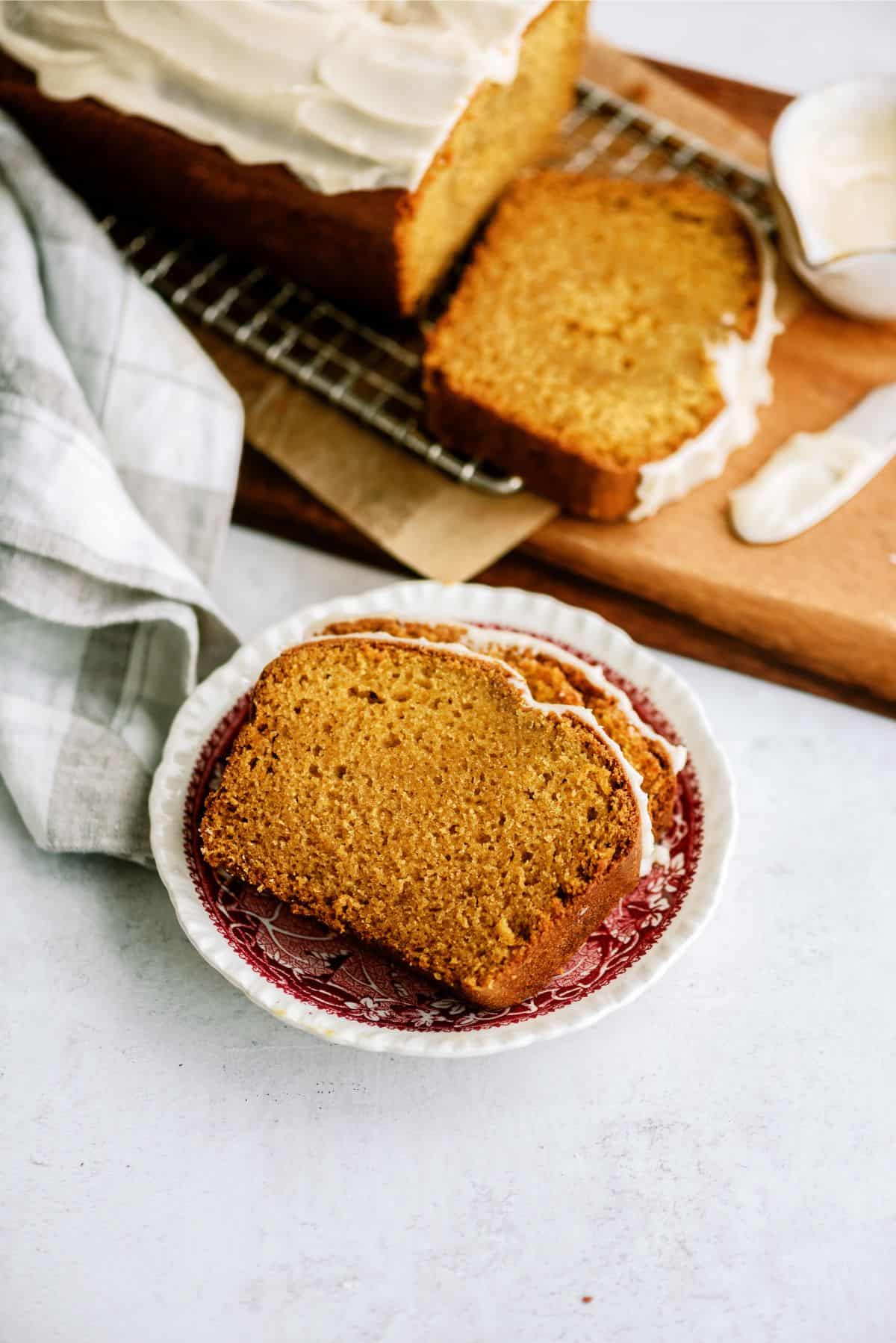 Two slices of frosted loaf cake on a red and white plate, with a loaf and more slices on a wooden board in the background. A checkered cloth sits to the left.