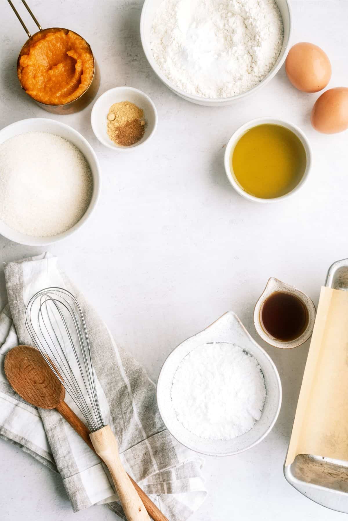 Ingredients for baking on a white background: flour, eggs, sugar, pumpkin puree, oil, spices, vanilla extract, a whisk, a wooden spoon, and a kitchen towel.