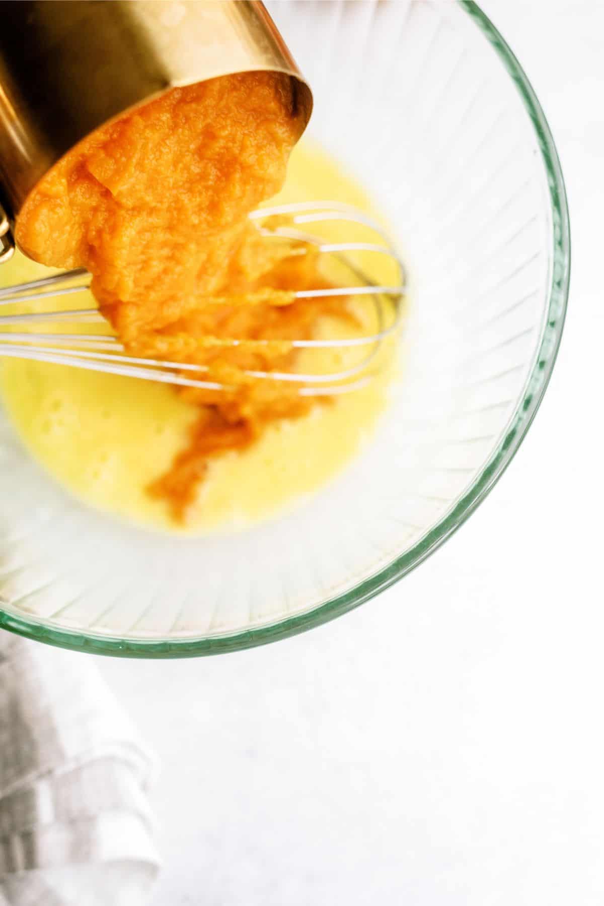 A cup of orange puree is being poured into a bowl containing yellow liquid, with a whisk inside the bowl, on a light-colored surface.