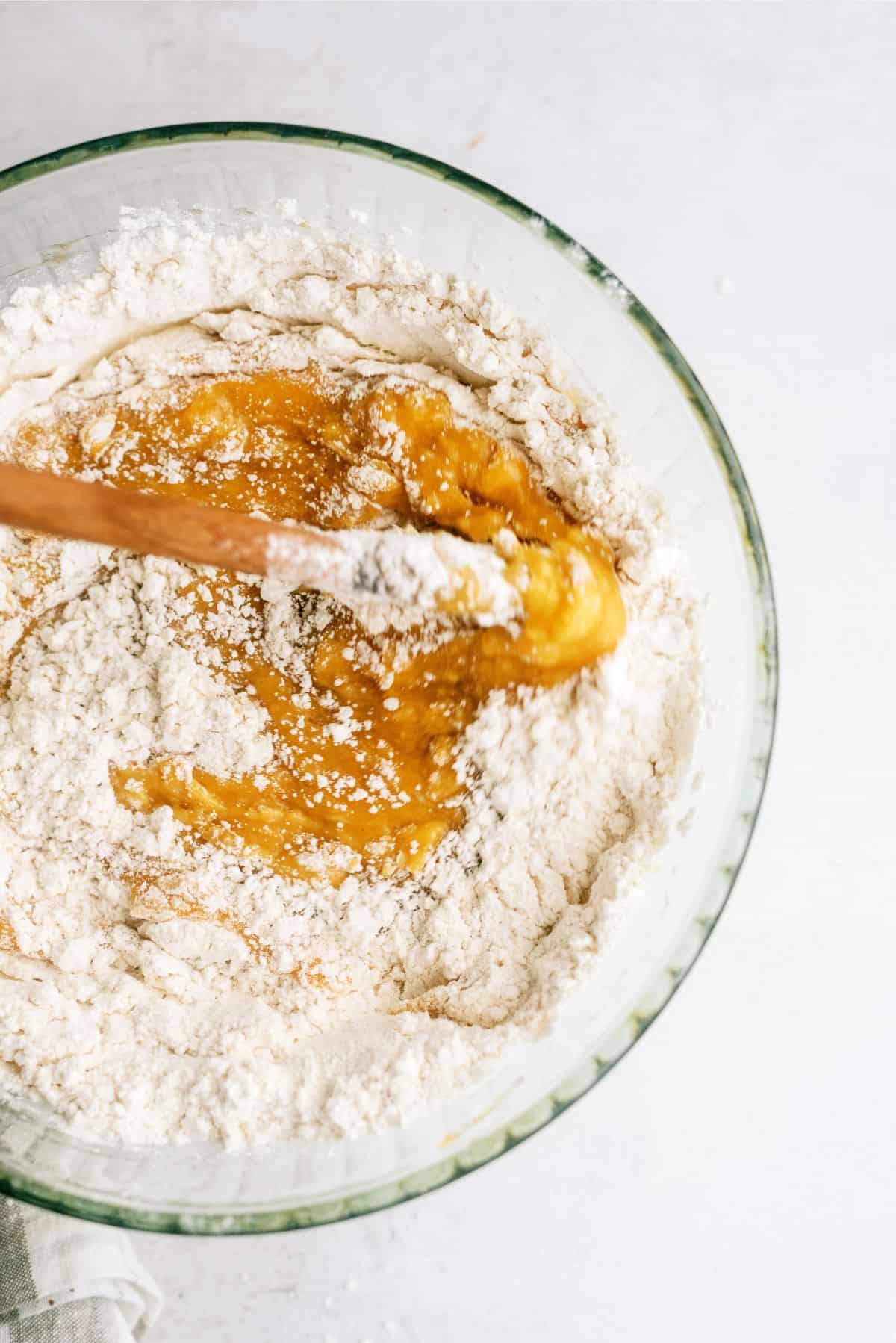 A wooden spoon mixes yellow batter with flour in a glass bowl on a light-colored surface.
