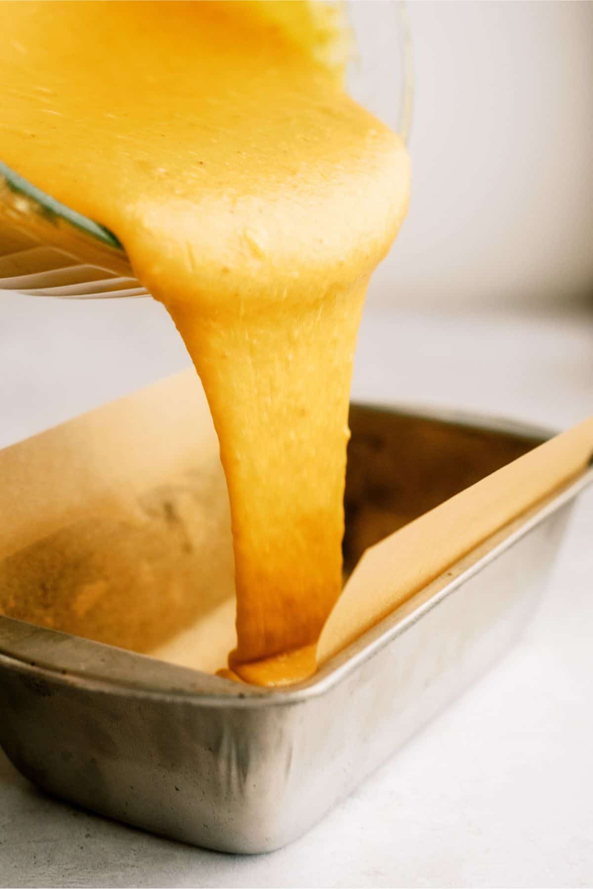 Liquid cake batter being poured from a glass bowl into a parchment-lined metal baking pan.