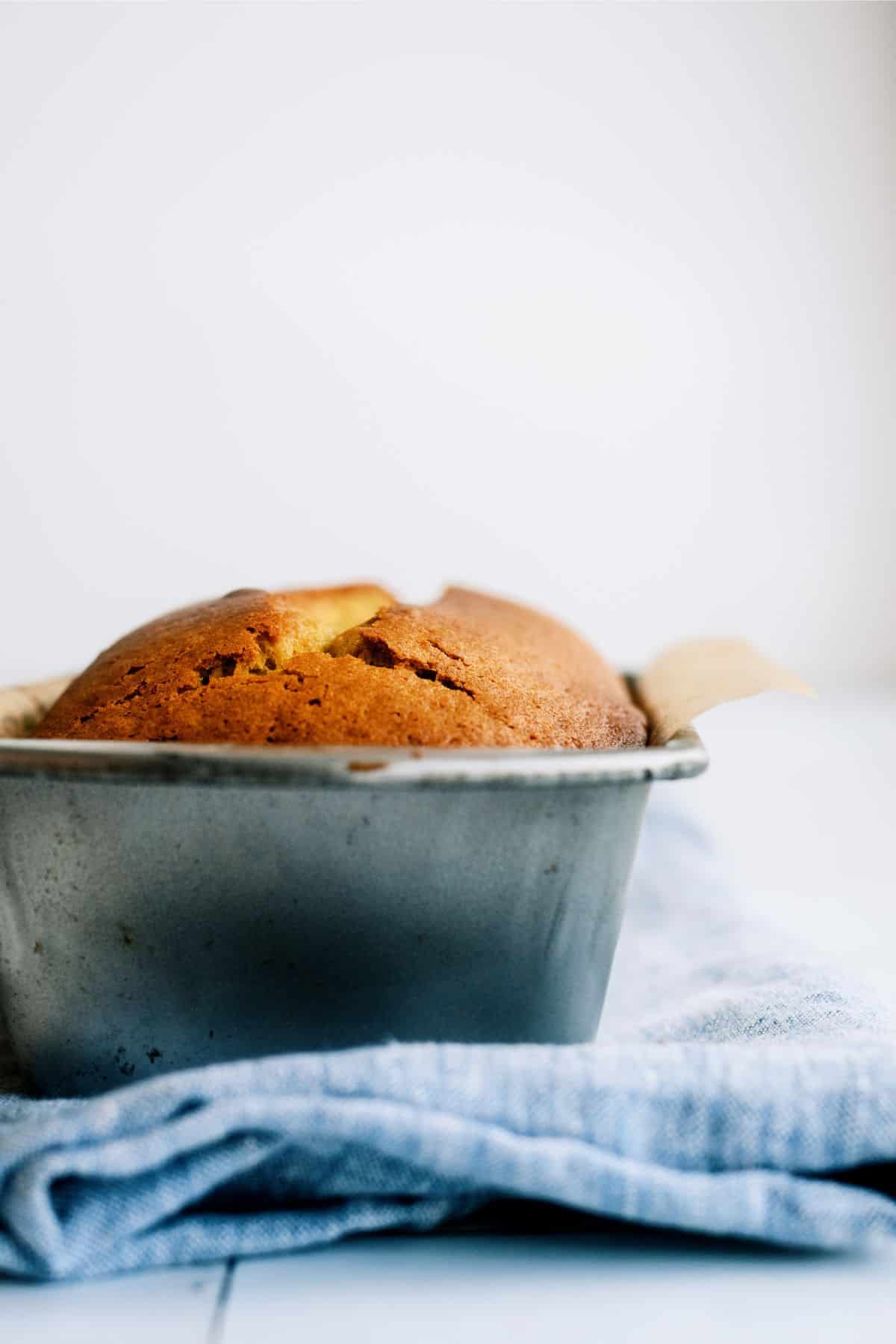 A single loaf of golden-brown bread in a metal baking tin rests on a blue kitchen towel.