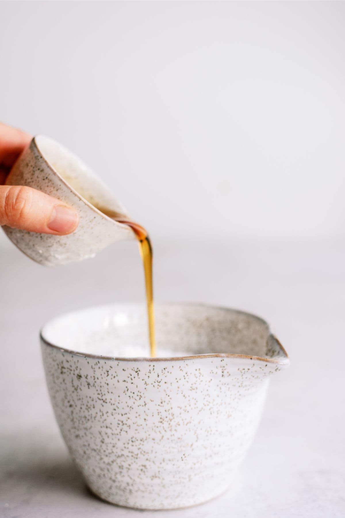 A person pours liquid from a small ceramic cup into a larger speckled ceramic bowl.
