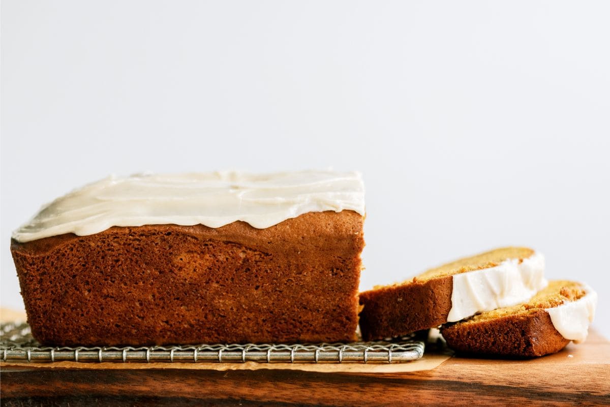 A loaf of frosted bread with two slices cut, placed on a cooling rack over a wooden surface.