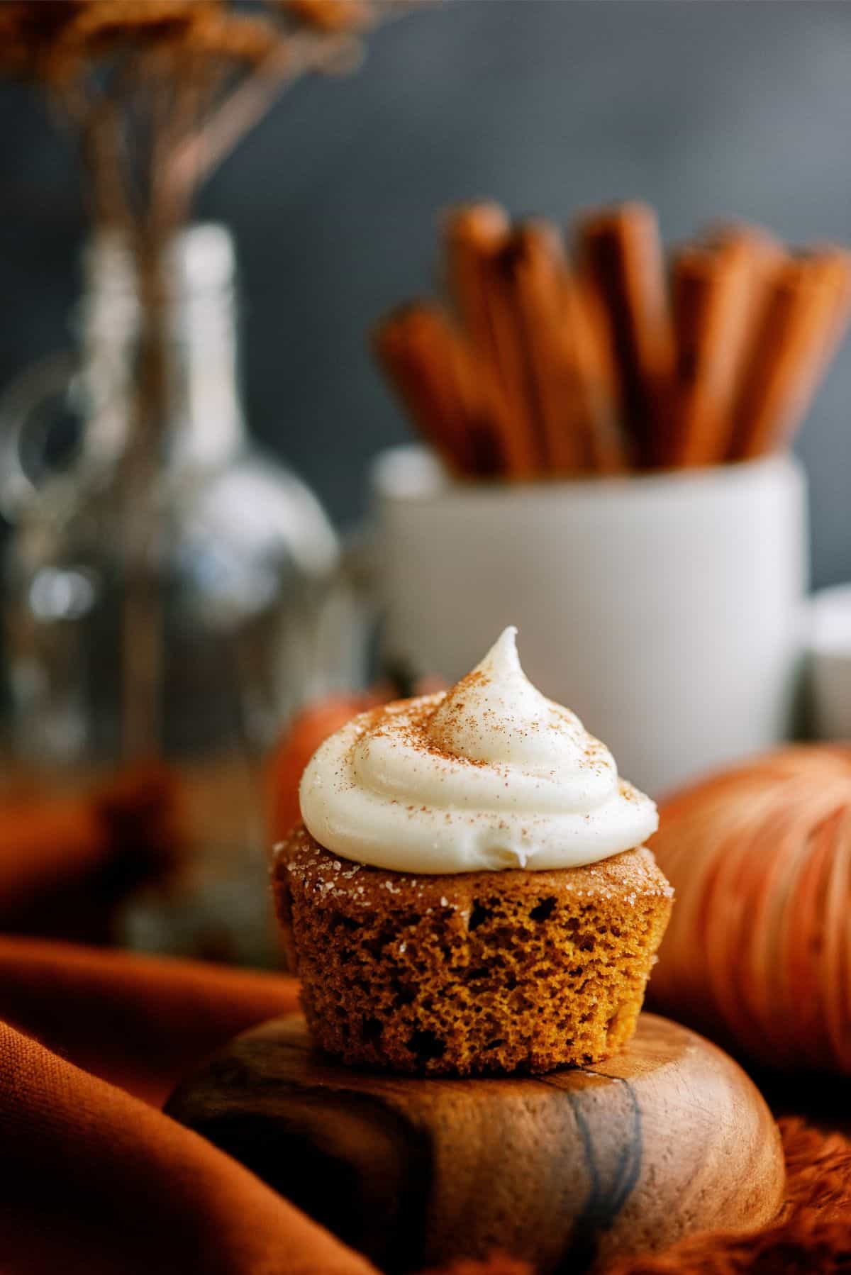 A pumpkin spice cupcake with cream cheese frosting on a wooden surface, surrounded by autumn-themed decorations including a jar and a bowl with cinnamon sticks in the background.