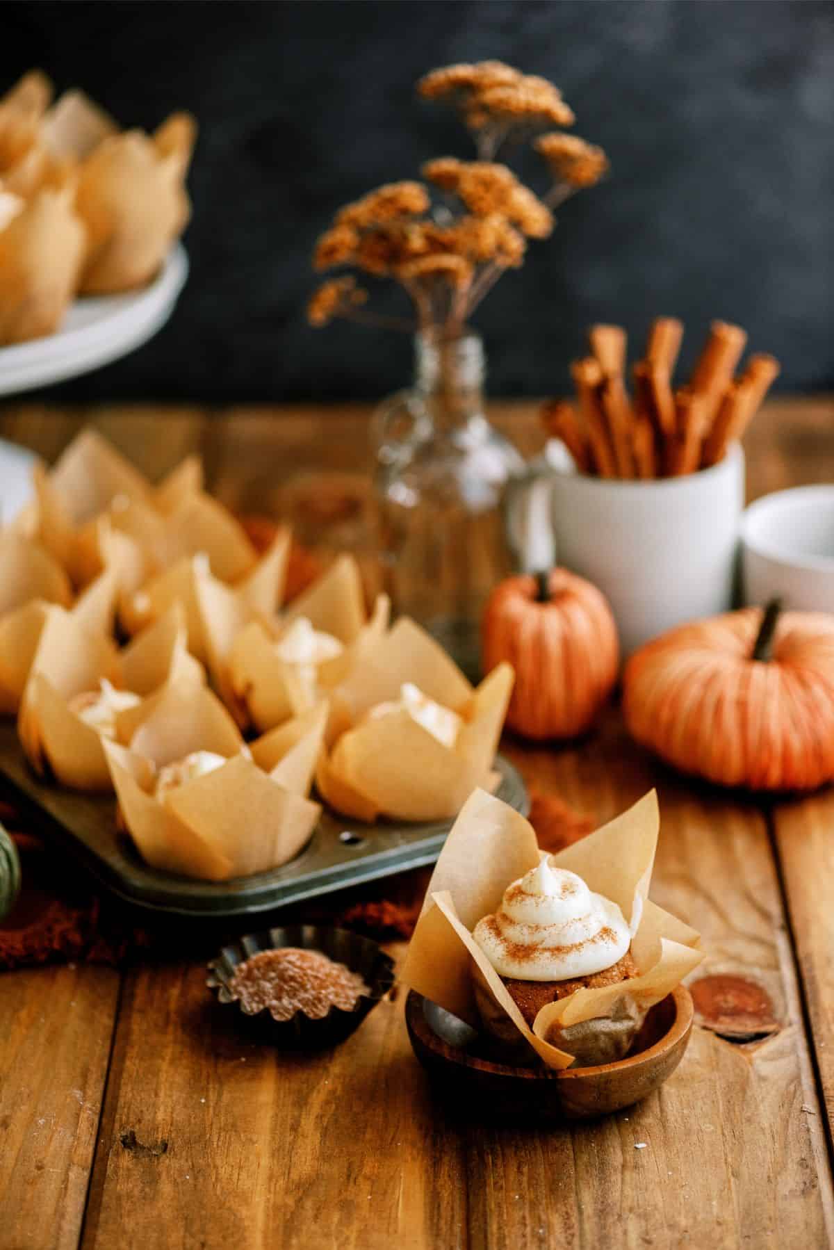 An arrangement of pumpkin muffins with cream cheese frosting in brown parchment cups on a wooden table with decorative pumpkins, a jar of cinnamon sticks, and dried flowers in a vase.