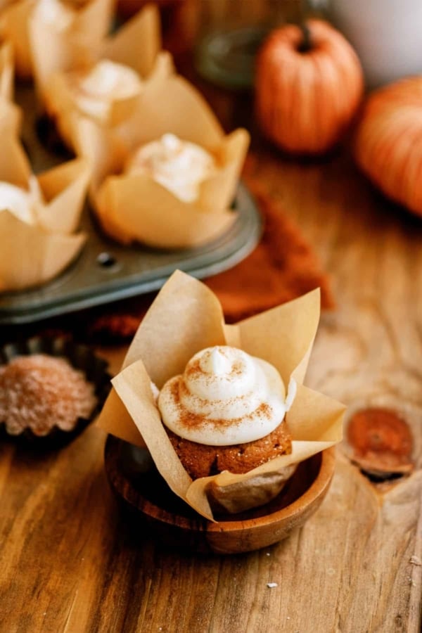 A pumpkin cupcake with cream cheese frosting, baked in a brown paper liner, is displayed on a wooden surface. In the background, a muffin tin with more cupcakes and small decorative pumpkins are visible.