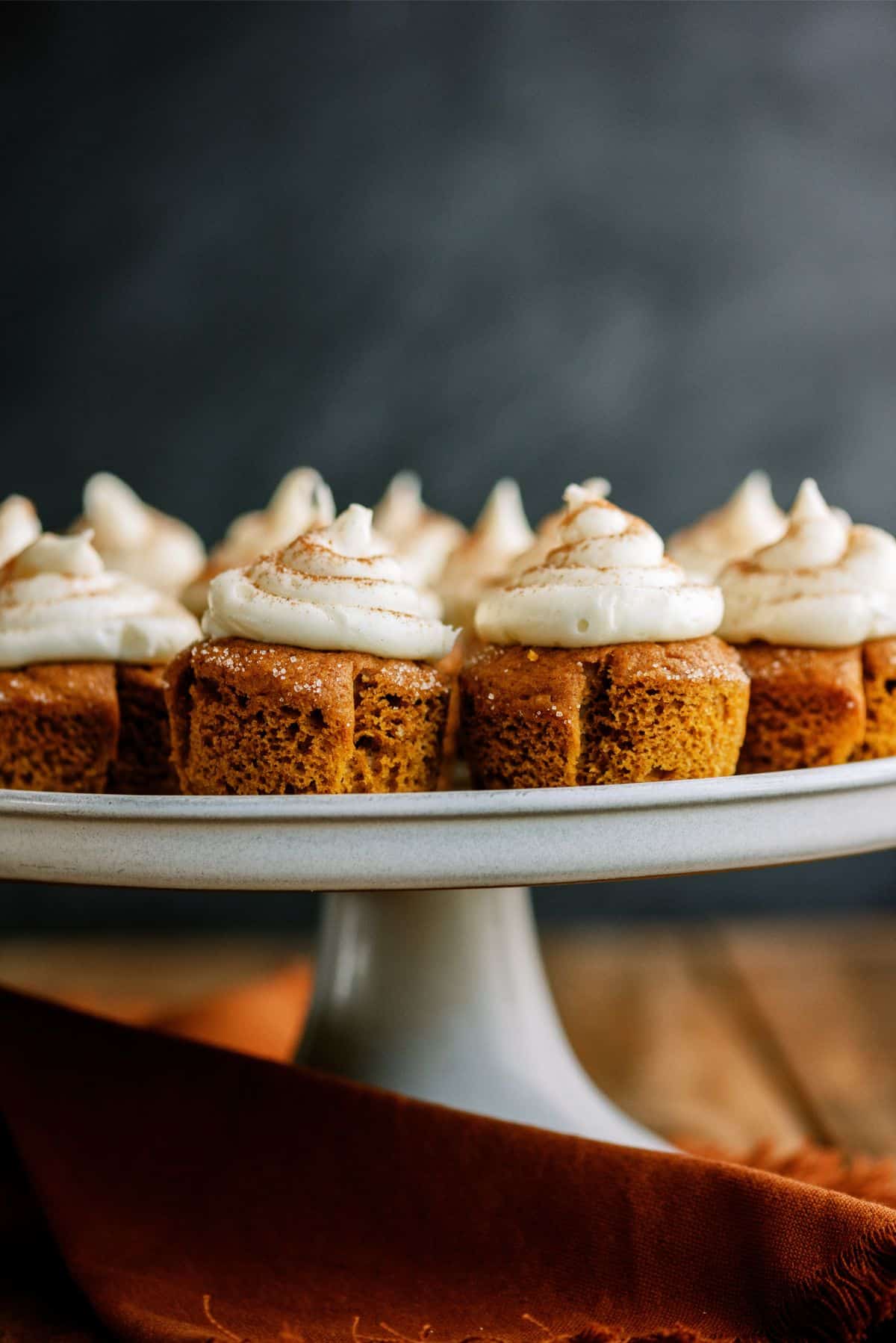 A cake stand holds multiple small square pieces of frosted pumpkin cake, each topped with a swirl of cream cheese icing and a sprinkle of cinnamon.