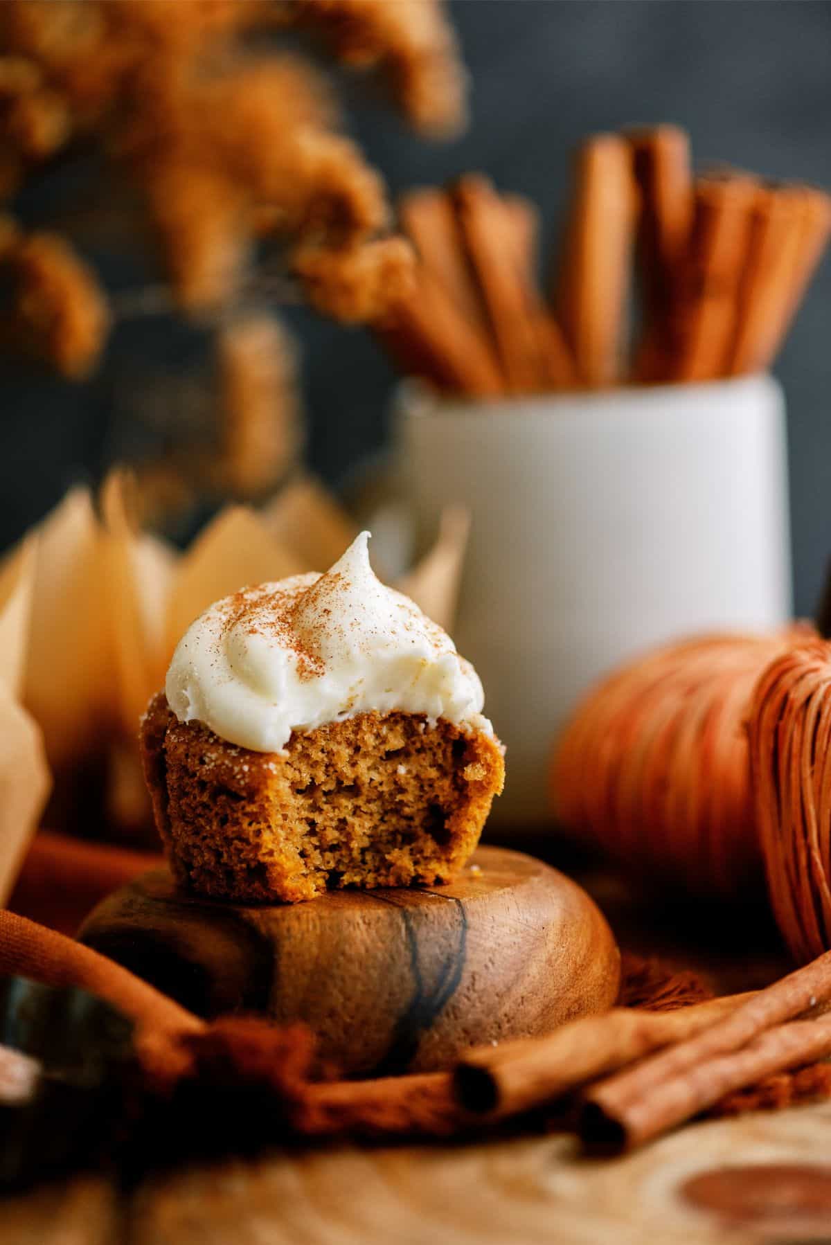A partially eaten cupcake with white frosting sits on a wooden surface surrounded by cinnamon sticks and autumn decor.