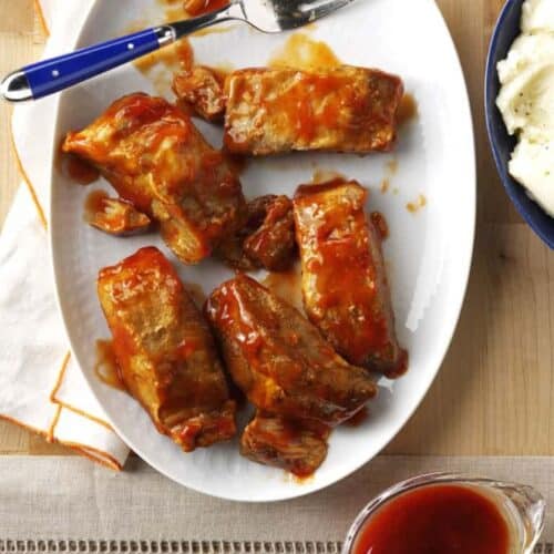 Five pieces of boneless ribs with barbecue sauce on a white oval plate, next to a fork, mashed potatoes, and a small pitcher of extra sauce.