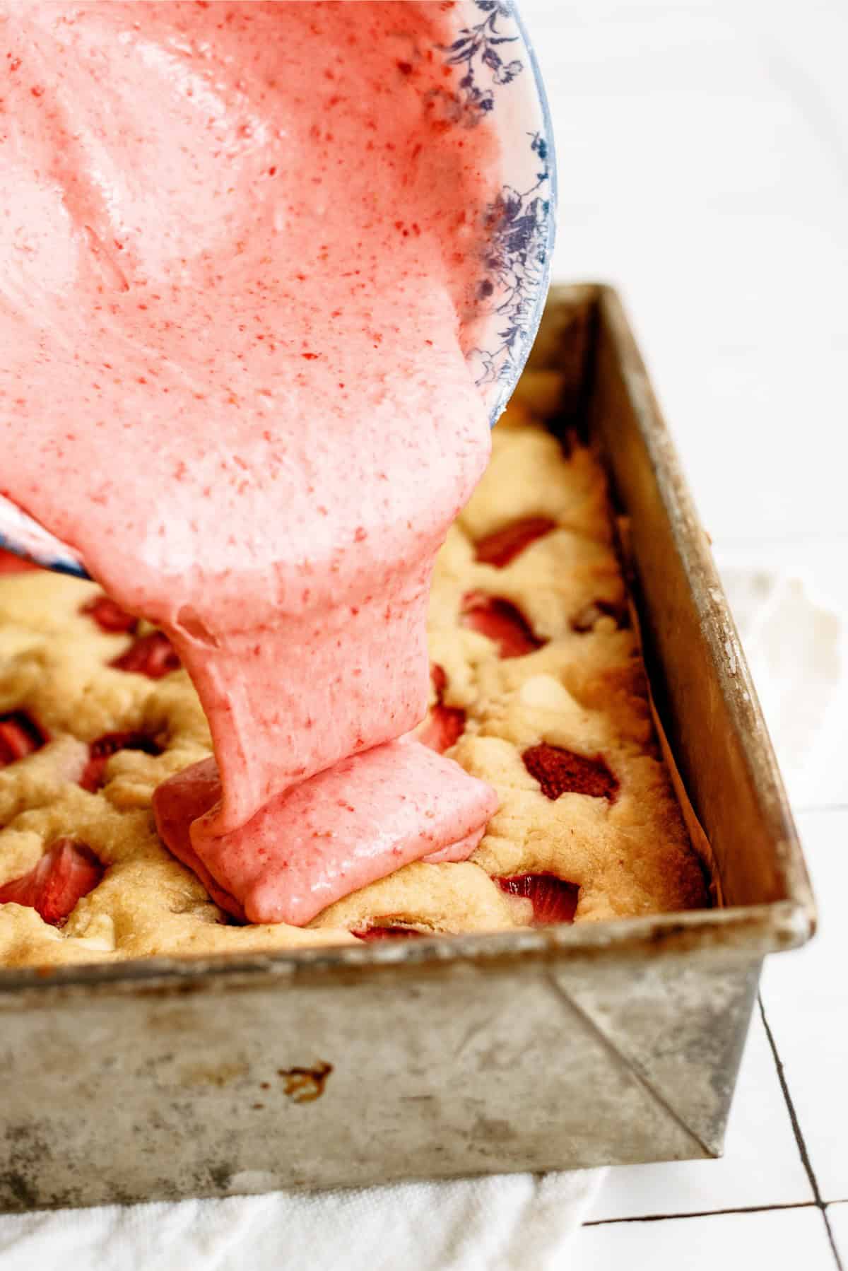 Strawberry frosting being poured on top of a pan with a strawberry blondies.