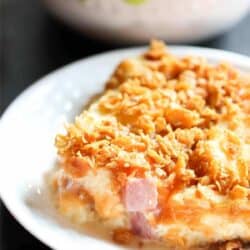 A serving of casserole with a crunchy topping on a white plate, with a bowl of green grapes in the background.