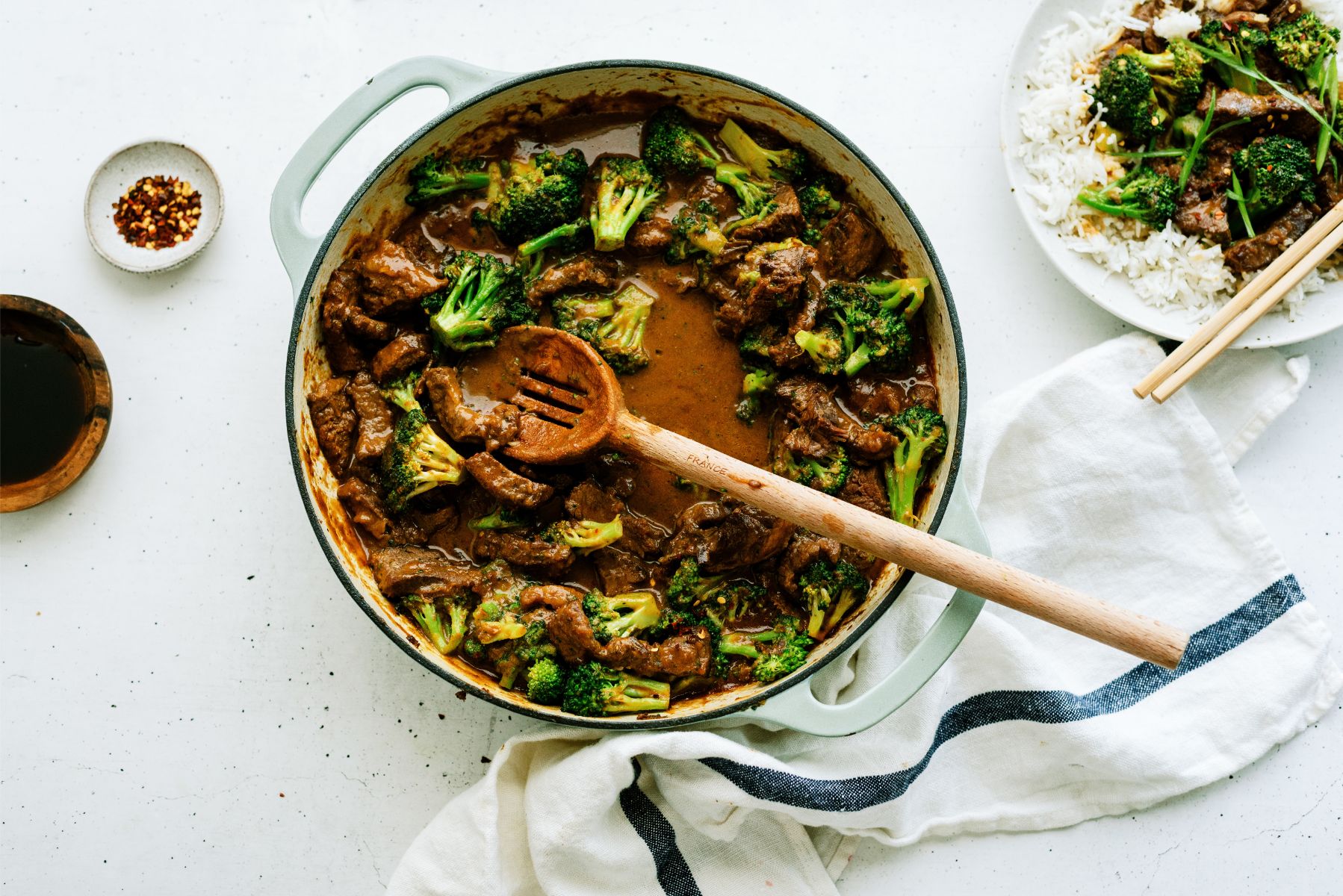 A pan of Beef and Broccoli with a plate on the side of rice, topped with Beef and Broccoli.