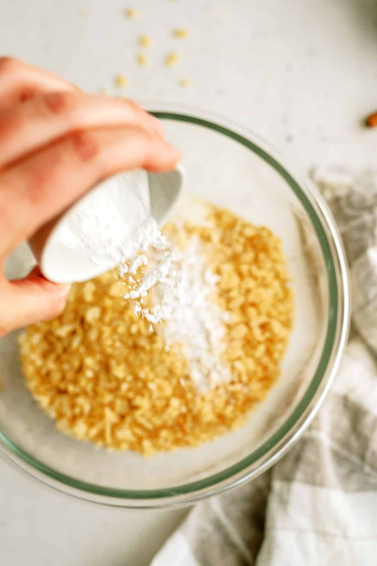 A glass mixing bowl with crushed cereal and a hand pouring a small cup of sugar, baking soda and salt into the bowl.