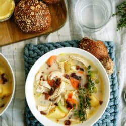 A bowl of creamy chicken soup with vegetables and herbs, served with whole grain rolls and a glass of water on a checkered tablecloth.