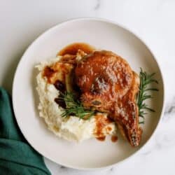 A plate with mashed potatoes, a pork chop topped with brown gravy, and a sprig of rosemary, on a white surface next to a green napkin.